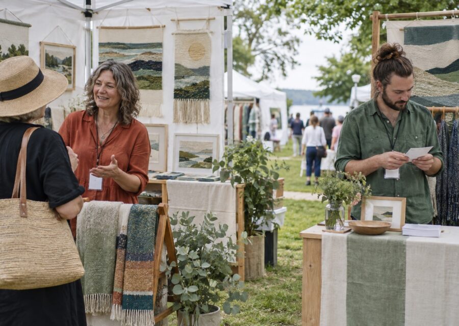Two textile artists dividing visitor and counter duties in an outdoor art fair booth.
